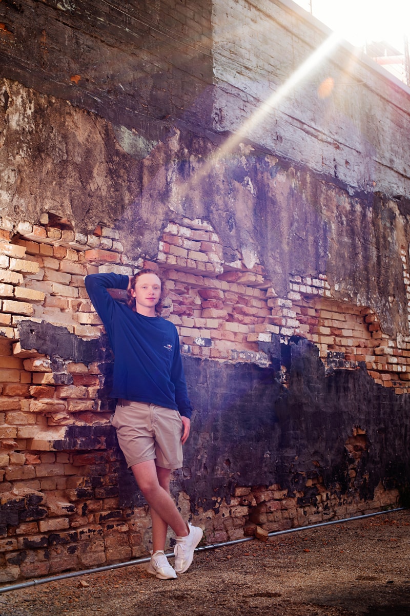 Senior Photographer, high school boy leans against an old brick wall confidently