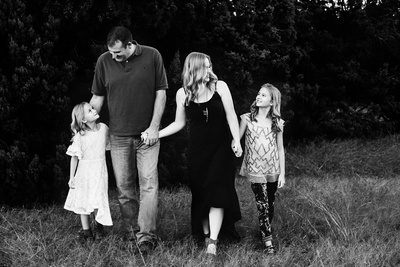 Family Photographer, father and mother hold hands with their two daughters as the walk in the meadow under trees