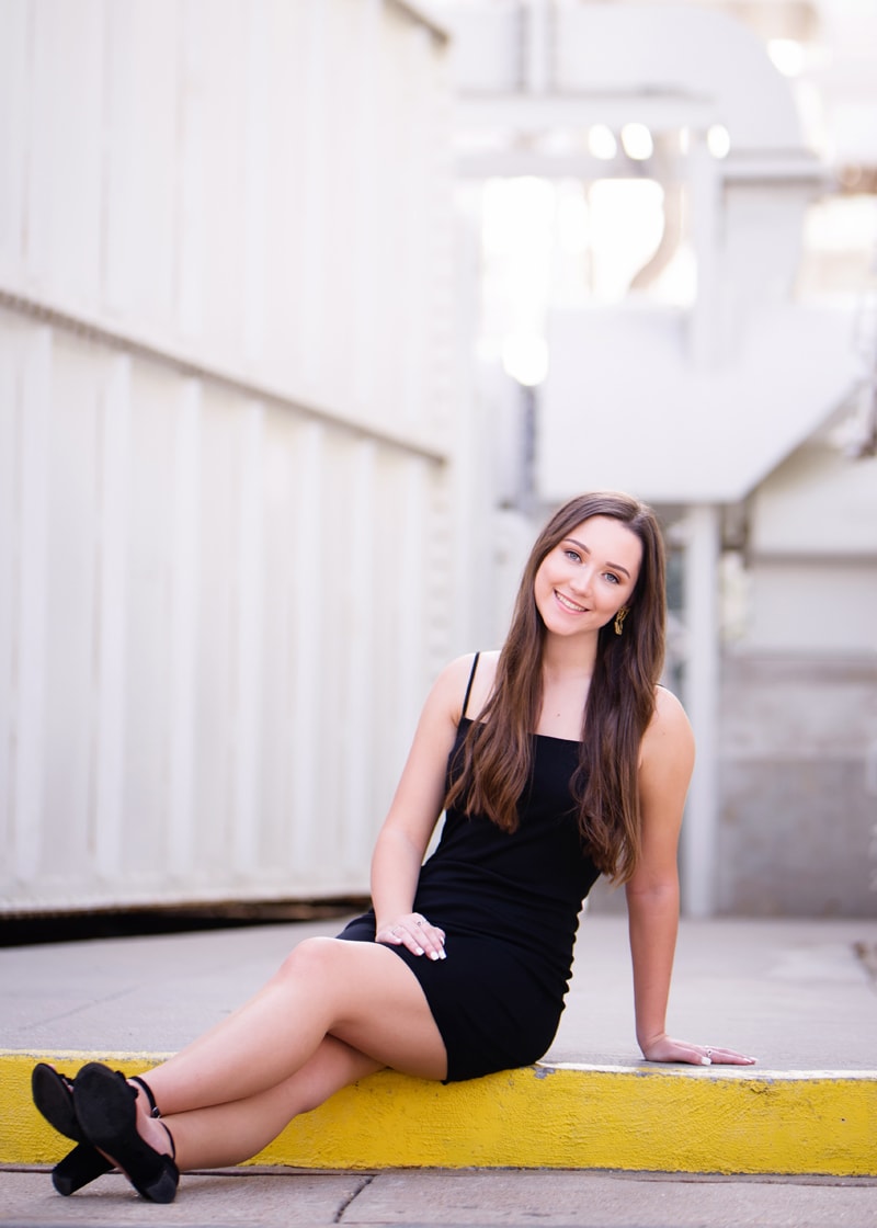 Senior Photographer, a high school girl sits on a curb, she is all dressed up and smiling