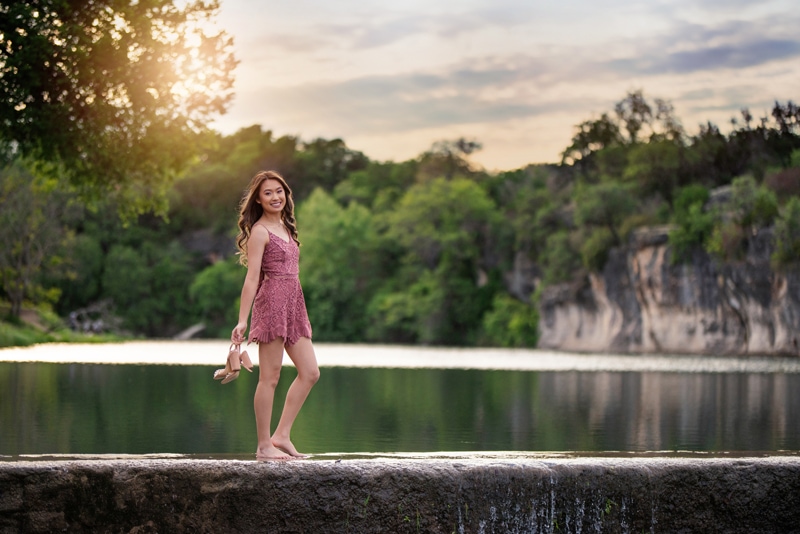 Senior Photographer, a high school woman takes off her shoes as she walks across a stone dam before quiet waters