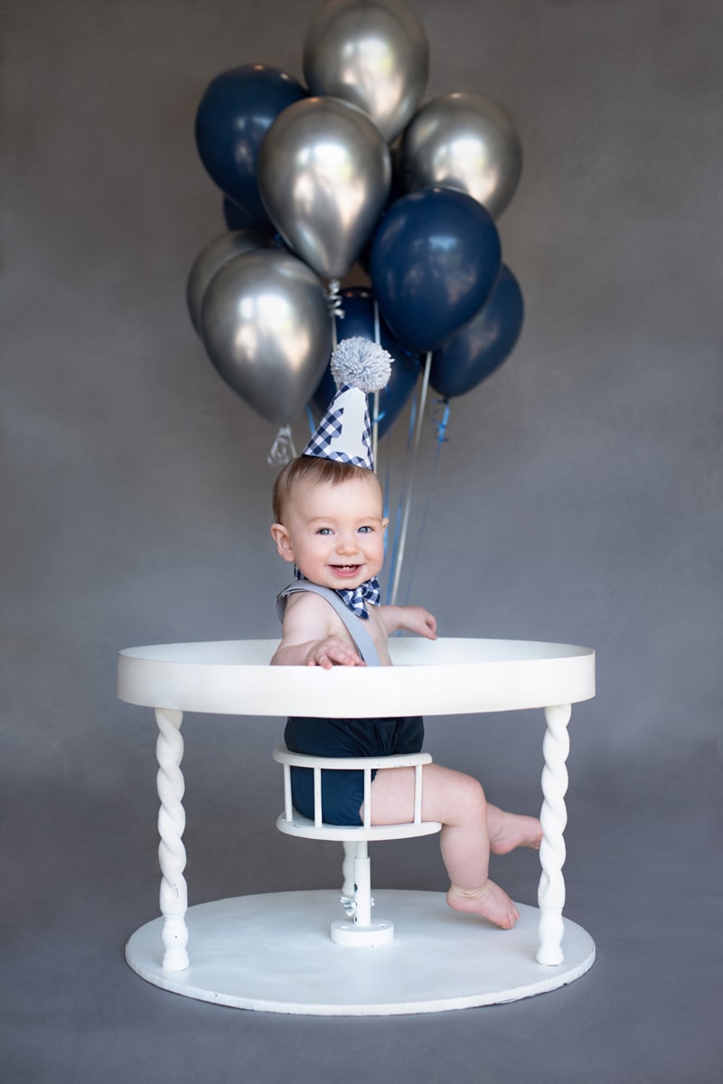 milestone photography, a baby boy sits in a vintage wooden chair and table, balloons above head.