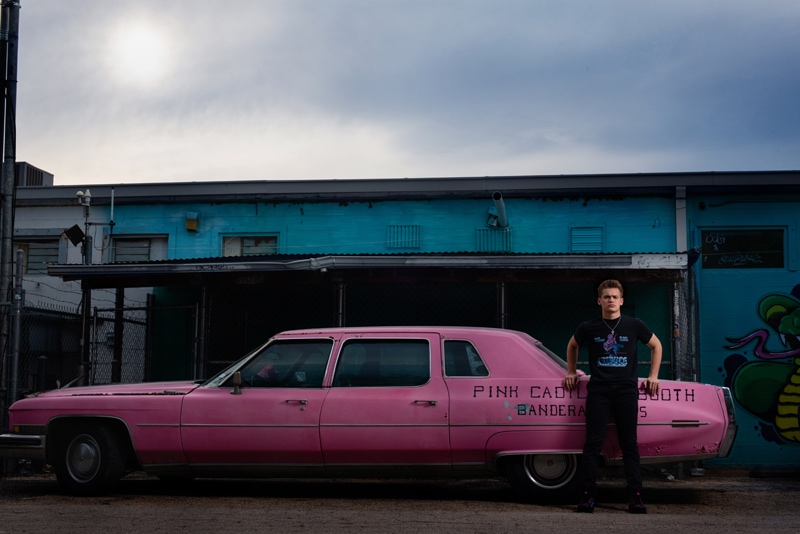 Senior Photographer, a young teenage man leans agains an old pink Cadillac