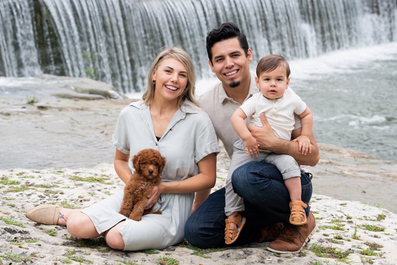milestone photography, a young father and mother sit with their baby son and puppy near a waterfall