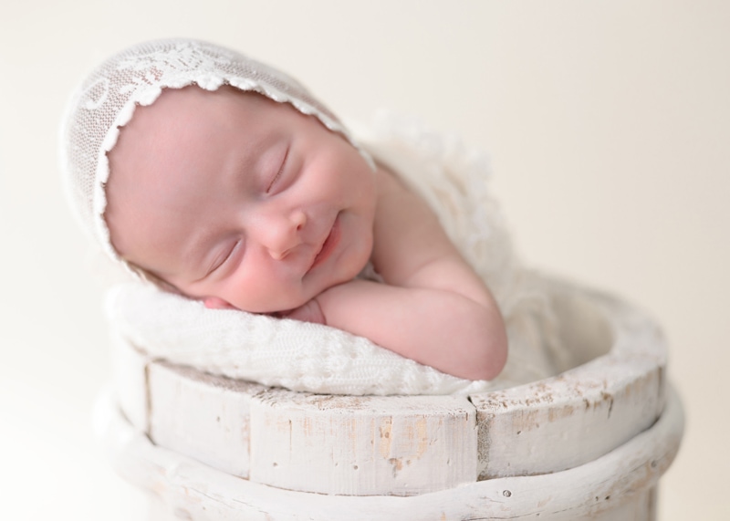 Newborn Photography, a baby smiles as she sleeps on a cushion within a wooden bucket