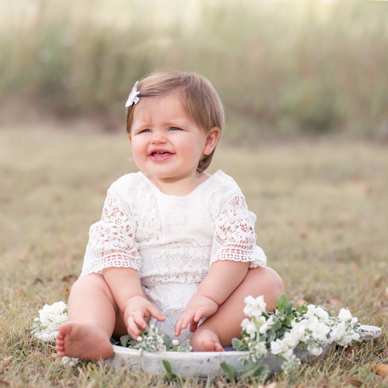 milestone photography, a baby girl sits on dry grass surrounded by flowers