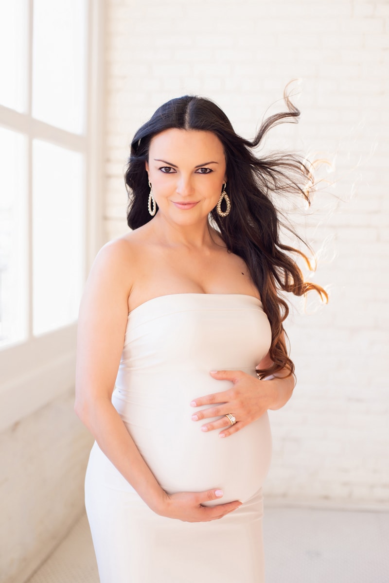 Maternity Photographer, pregnant woman holds belly in anticipation of baby, her hair blows into the air