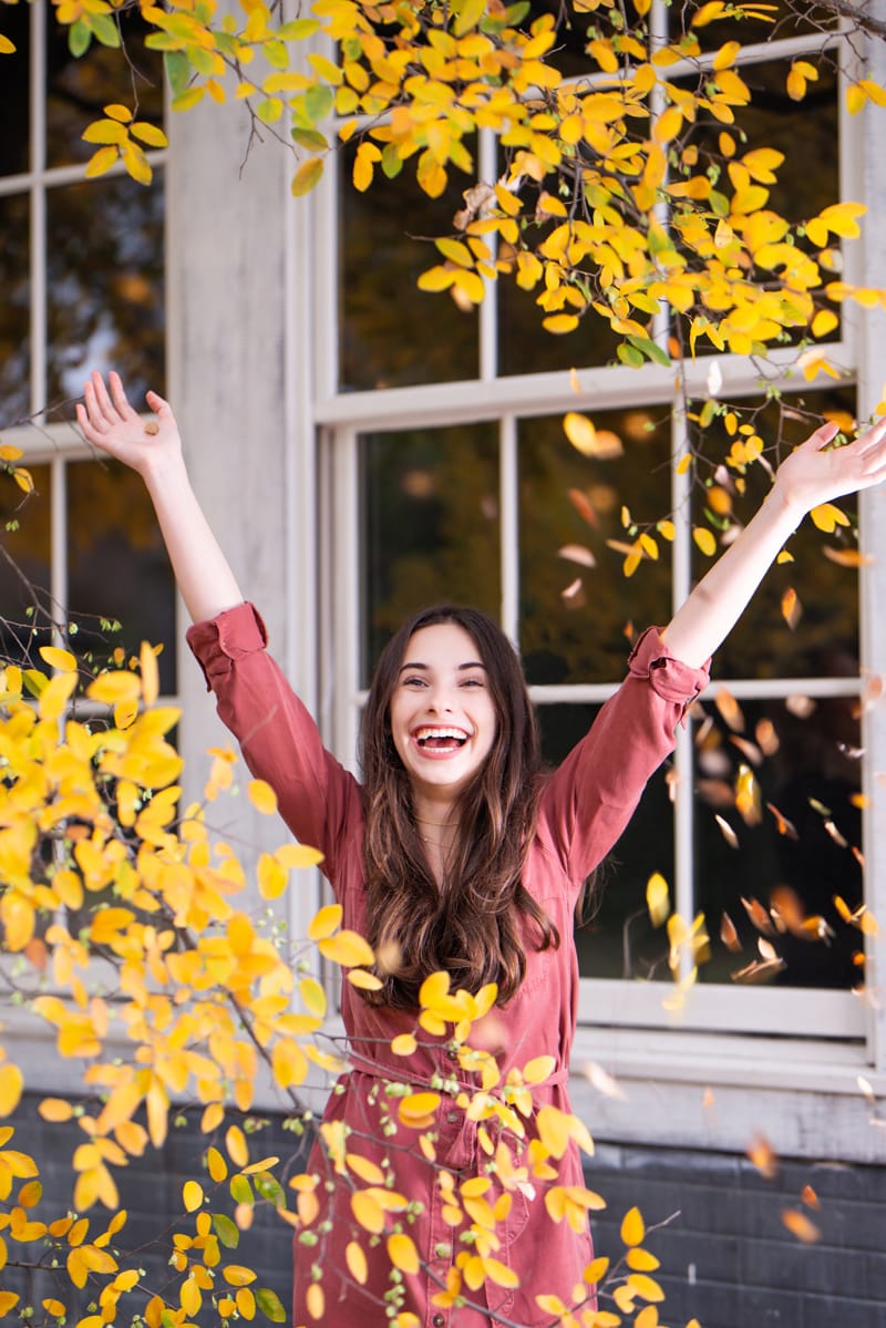 Senior Photographer, a highschool girl throws her hands in the hair amidst the fall leaves on a tree, it looks like golden confetti before her