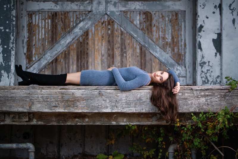 Senior Photographer, a young teenage woman lays on a large wooden beam near a farm yard
