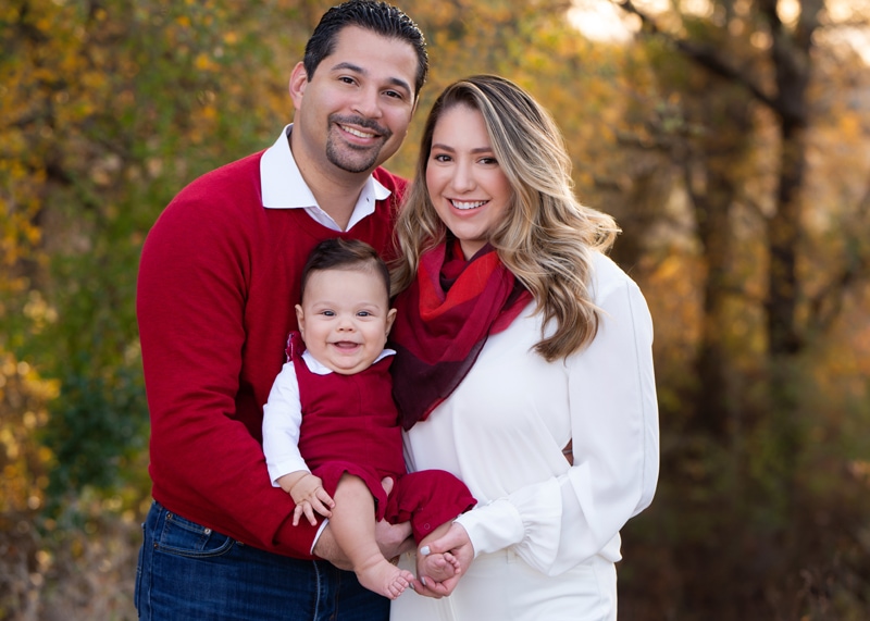 Family Photographer, young mom and dad stand smiling as they hold their happy baby in the woods