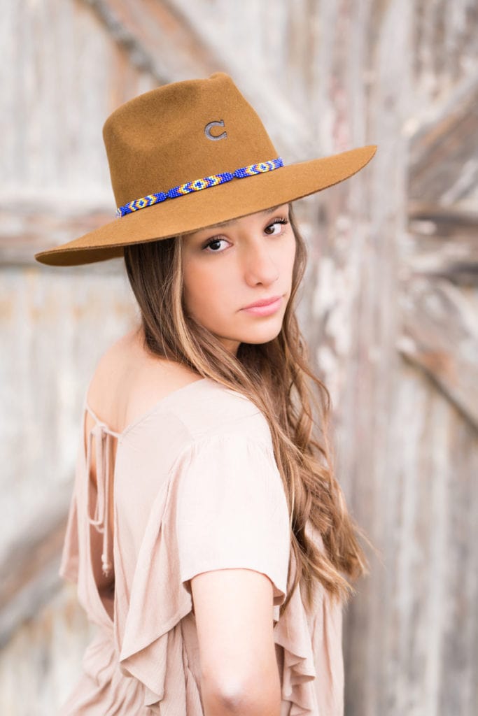 Senior Photographer, High school girl looks over her shoulder content, she wears a wide brim fedora and stands before barn doors