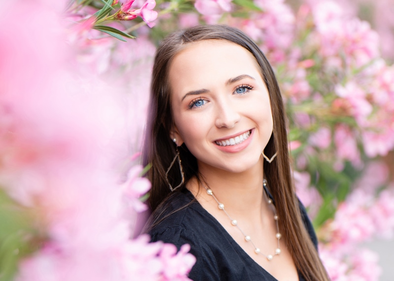 Senior Photographer, a high school woman wears jewelry and smiles amongst a large bush with pink flowers