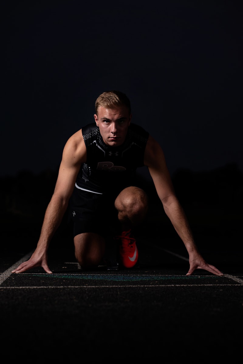 Senior Photographer, high school boy kneels ready to run, he wears track shoes and jersey