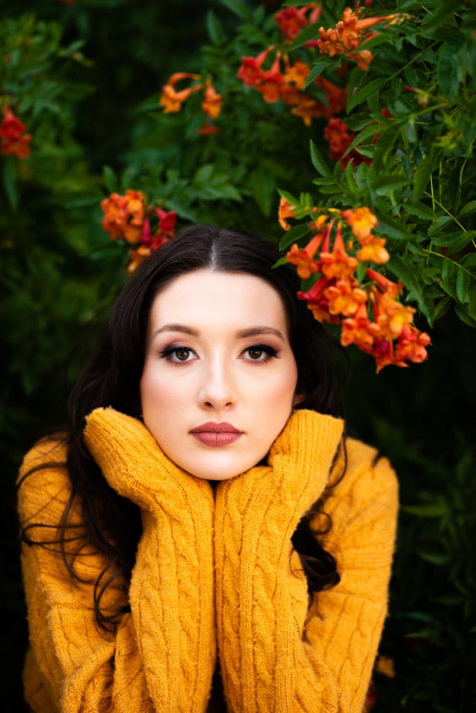 Senior Photographer, A high school woman leans in and holds her face up with her hands confidently, she stands before a vibrant green hedge with orange flowers