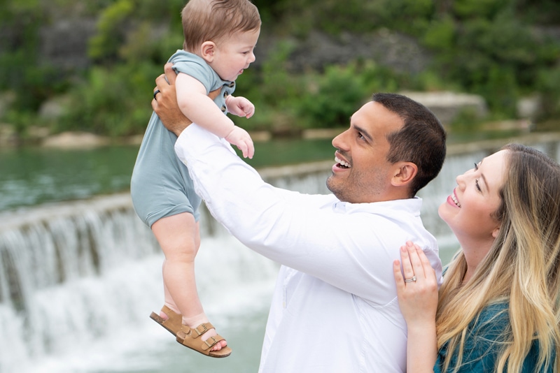 a mother and father look on to their baby, they stand before a waterfall, dad holds up baby