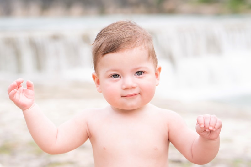 a baby boy lifts his arms as he stands before a waterfall outdoors