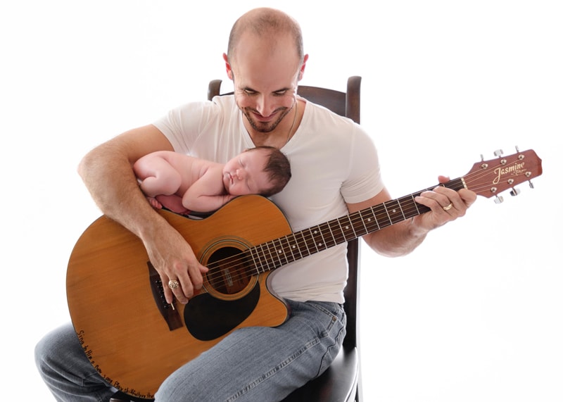 Newborn Photography, a father plays the guitar softly as baby lays on top of guitar sleeping