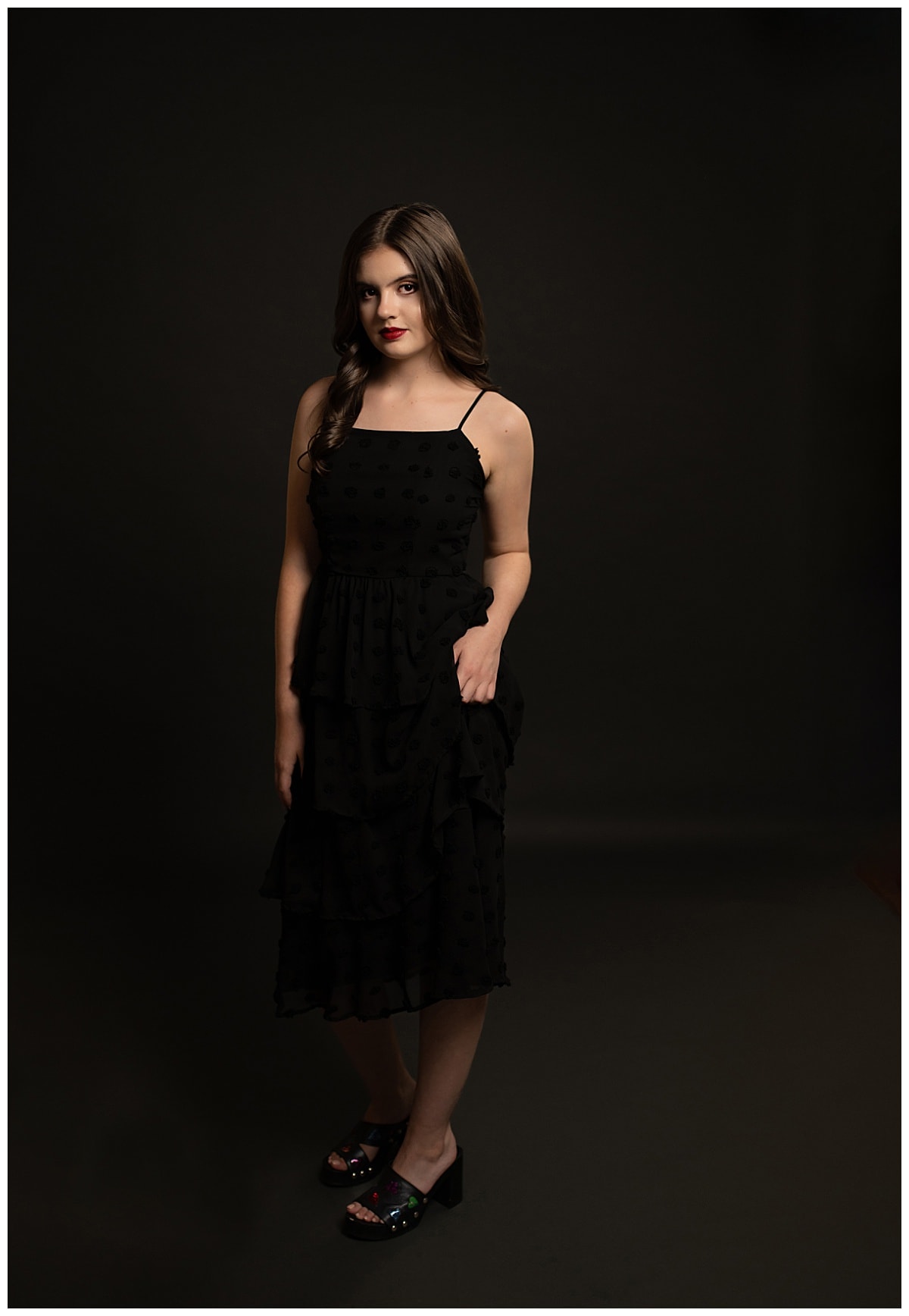 senior in black dress stands in front of dark background by Austin Senior Photographer